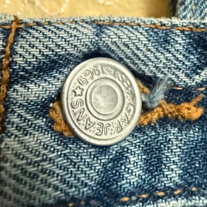 May include: Close-up of a silver metal button on blue denim jeans. The button is embossed with the words "GAP JEANS" and the number "696". The button is surrounded by a star and a circular design.