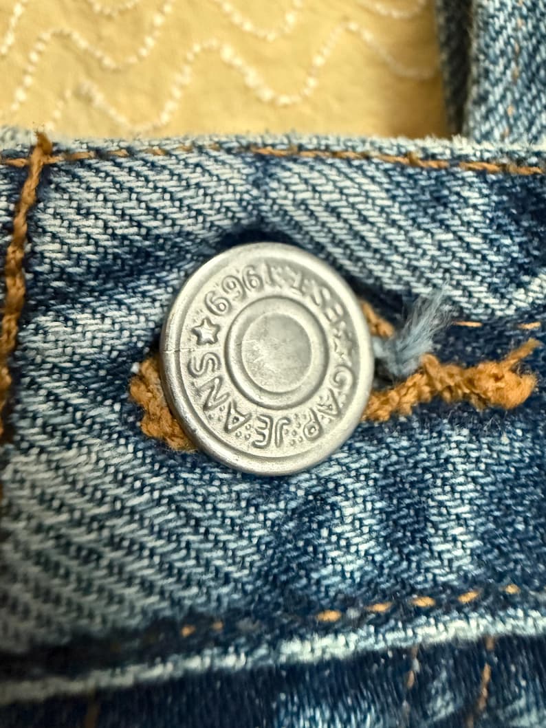 May include: Close-up of a silver metal button on blue denim jeans. The button is embossed with the words "GAP JEANS" and the number "696". The button is surrounded by a star and a circular design.