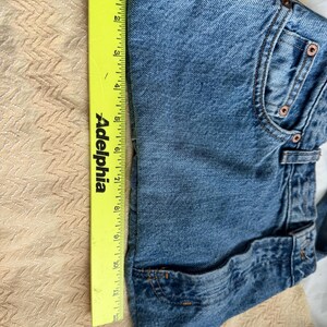 May include: Folded blue denim shorts with a yellow ruler alongside. The ruler shows a measurement of approximately 12 inches. The shorts have classic five-pocket styling and copper-colored rivets.