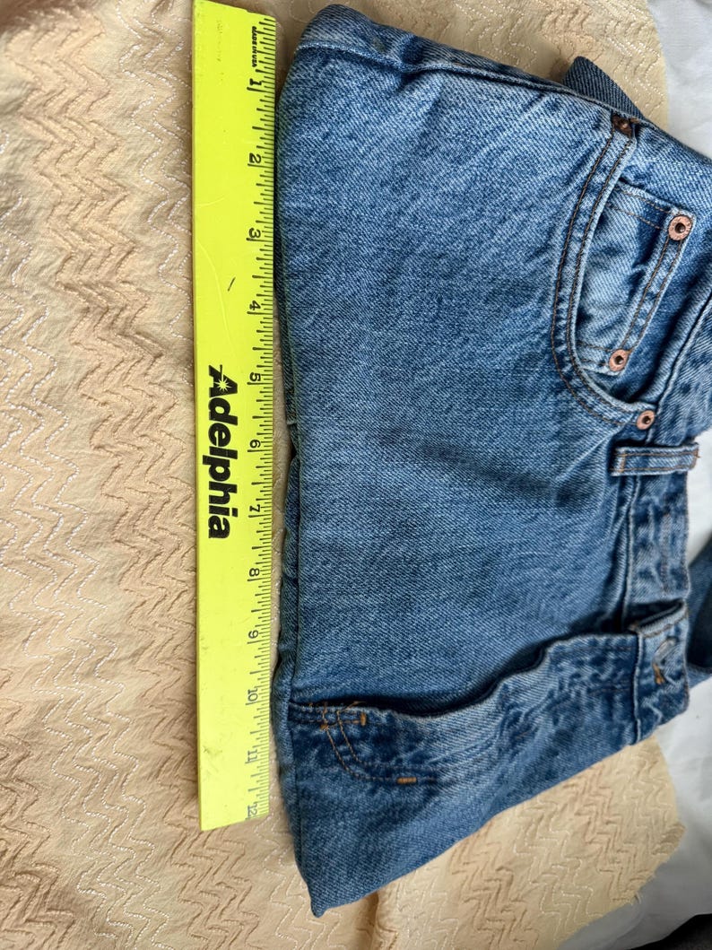 May include: Folded blue denim shorts with a yellow ruler alongside. The ruler shows a measurement of approximately 12 inches. The shorts have classic five-pocket styling and copper-colored rivets.