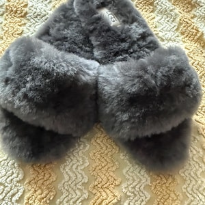 May include: A pair of dark gray open-toe slippers featuring a thick, plush, fuzzy texture across the top straps. The slippers have a slip-on design with a white rectangular label on the inner footbed that displays the brand name UGG in black text.