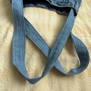 May include: A handmade denim tote bag, crafted from repurposed blue jeans. The bag features the original waistband and belt loops, with long straps made from the jean fabric. The bag is shown against a textured, light-colored background.