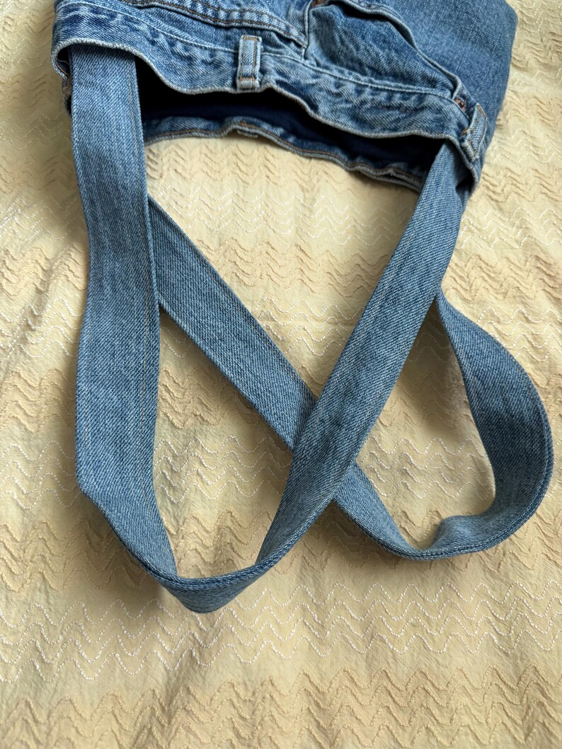 May include: A handmade denim tote bag, crafted from repurposed blue jeans. The bag features the original waistband and belt loops, with long straps made from the jean fabric. The bag is shown against a textured, light-colored background.