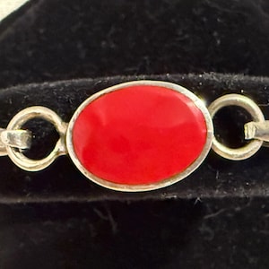 May include: Silver bracelet featuring a vibrant red oval-shaped stone. The stone is set in a silver frame and connected to the bracelet with silver loops. The bracelet is displayed on a black velvet surface.