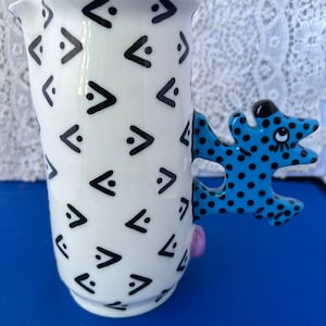 May include: White ceramic pitcher with a black geometric pattern and a blue and black polka dot handle shaped like a cartoon animal. The handle has a purple accent.