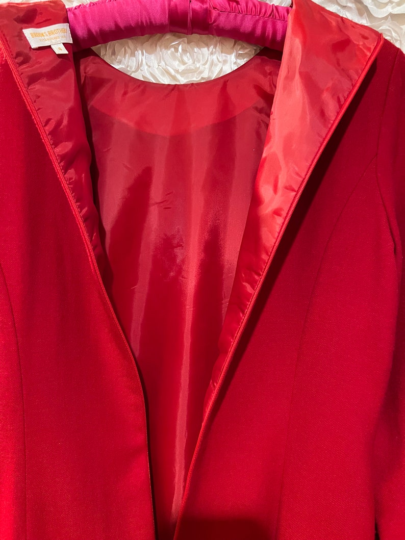 Puede incluir: Un blazer rojo con dise&ntilde;o de cuello en V, colgado de una percha rosa. El forro interior es de un tono rojo m&aacute;s claro. El blazer es de la marca Brooks Brothers.