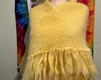 May include: A bright yellow knitted shawl with a fringed bottom. The shawl is displayed on a mannequin. The background features a tie-dye design with the word "PEACE" in white letters.