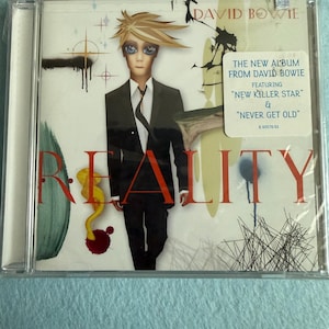 May include: A CD case featuring the album "Reality" by David Bowie. The cover art depicts a stylized illustration of Bowie in a black suit. The album title is in large red letters, with the text "The New Album From David Bowie" and song titles also visible.