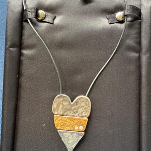 May include: A silver and gold-toned heart pendant necklace with the words "wisdom", "love", and "faith" inscribed. The pendant has a black cord and is displayed on a black satin surface. The pendant has three small clear stones.