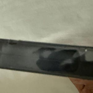 May include: A black plastic DVD case spine viewed from a side angle. The surface of the black plastic is smooth and reflects light, showing a dark, indistinct shape or shadow on the center portion of the spine. The edges are straight and defined.