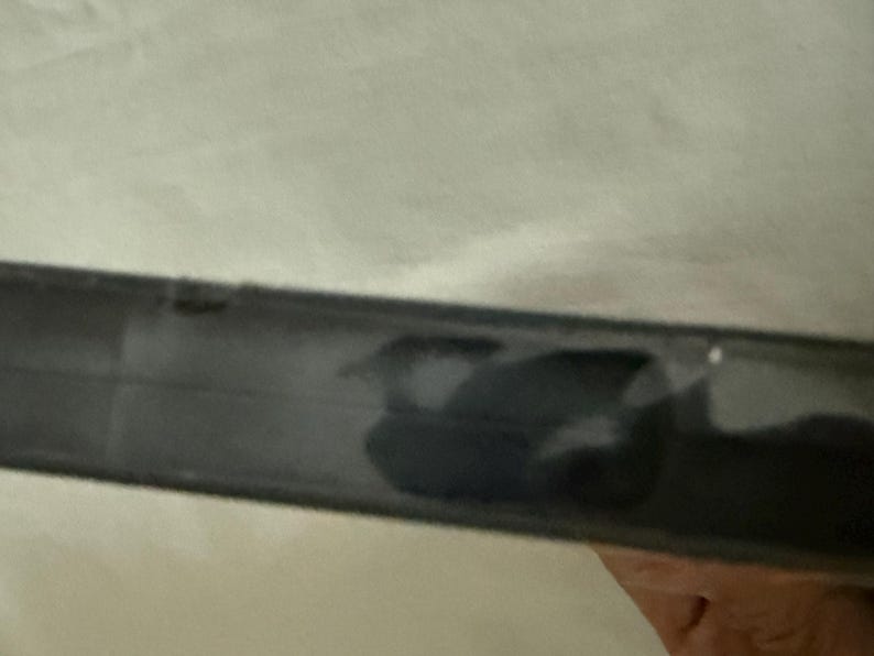May include: A black plastic DVD case spine viewed from a side angle. The surface of the black plastic is smooth and reflects light, showing a dark, indistinct shape or shadow on the center portion of the spine. The edges are straight and defined.