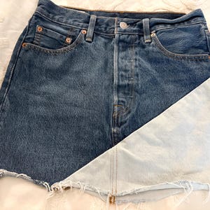 May include: A denim skirt with a unique design. The skirt is primarily blue denim, with a light blue panel on the right side. It has a button closure and classic five-pocket styling. The hem is frayed, adding a casual touch to the garment.