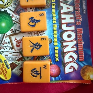 May include: Four orange Mahjong tiles with blue symbols and letters N, S, E, and W are displayed. The game box reads "Moraff's Maximum Mahjongg" and "4 Complete Mahjongg Games". The background includes game pieces and text.
