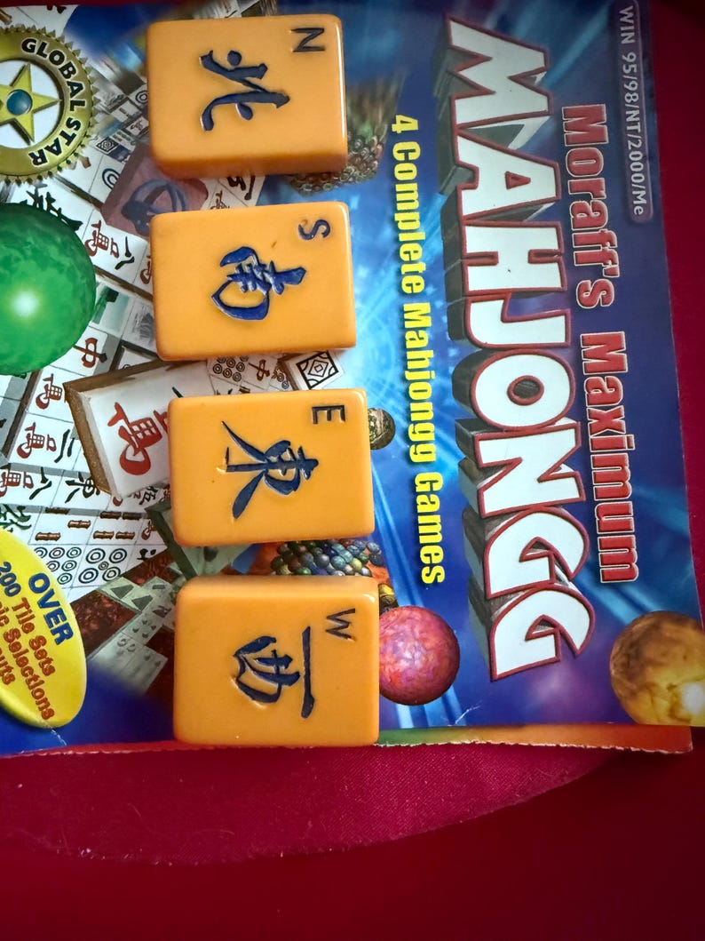 May include: Four orange Mahjong tiles with blue symbols and letters N, S, E, and W are displayed. The game box reads "Moraff's Maximum Mahjongg" and "4 Complete Mahjongg Games". The background includes game pieces and text.