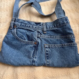 May include: A blue denim shoulder bag, crafted from repurposed jeans. The bag features the original pockets, belt loops, and a denim strap. The bag is a light blue wash, with visible stitching and rivets, showcasing a unique, upcycled design.