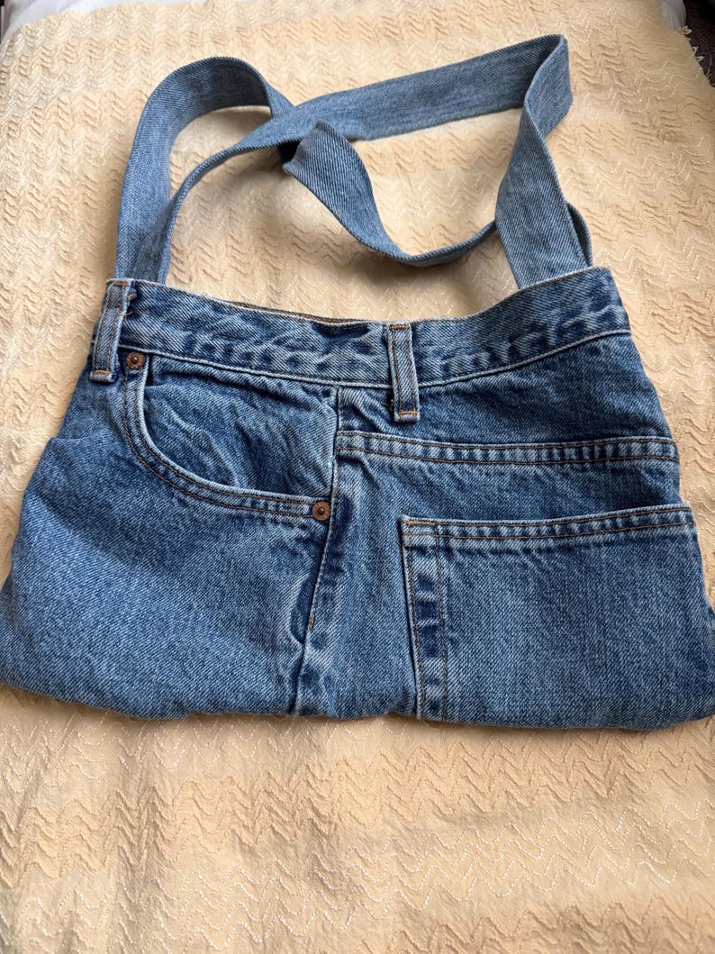 May include: A blue denim shoulder bag, crafted from repurposed jeans. The bag features the original pockets, belt loops, and a denim strap. The bag is a light blue wash, with visible stitching and rivets, showcasing a unique, upcycled design.