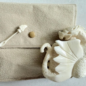 May include: A cream-colored dragon figurine with detailed scales and wings, accompanied by a small matching spoon, resting next to a beige suede-like pouch featuring a single snap button closure. The set is monochromatic and textured.