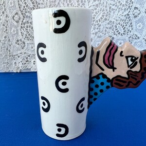 May include: White ceramic mug with a black and white geometric pattern. The mug has a unique handle shaped like a cartoon face with brown hair, blue eyes, and a pink tongue.