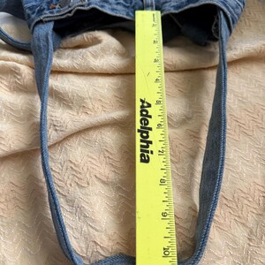 May include: A light blue denim tote bag with long straps. The bag is made from recycled denim and has a measurement of approximately 10 inches in height. The bag is on a light peach colored surface.