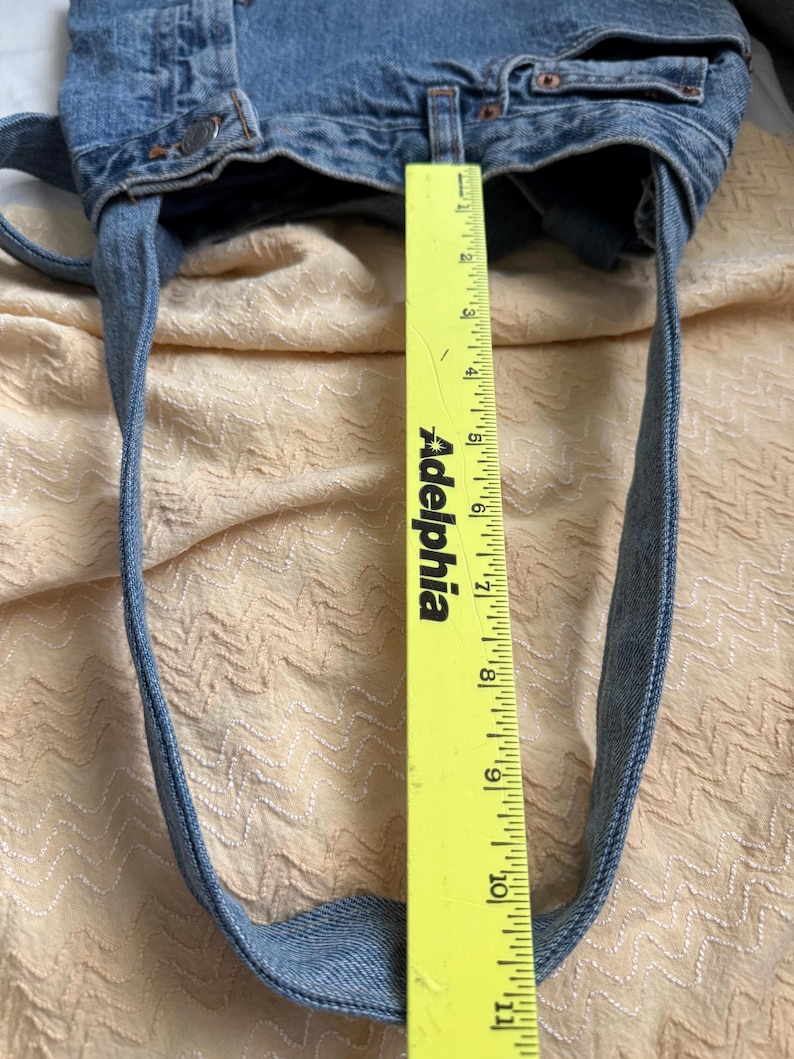 May include: A light blue denim tote bag with long straps. The bag is made from recycled denim and has a measurement of approximately 10 inches in height. The bag is on a light peach colored surface.