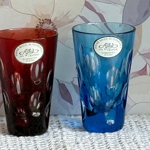 May include: Two colored crystal drinking glasses. One is a deep red, the other is a vibrant blue. Both glasses have a textured pattern and a label that reads "Hand Made in Hungary". The glasses are on a white, textured surface.
