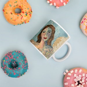 Może przedstawiać: Biały kubek ceramiczny z kolorową ilustracją twarzy kobiety na niebieskim tle. Kubek ma uchwyt po prawej stronie. Kubek jest otoczony pączkami z kolorowymi posypkami.