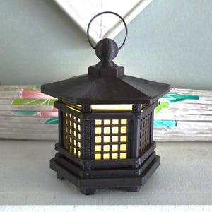 May include: A black, Japanese-style lantern with a square grid design on the sides. The lantern has a handle on top and emits a warm, yellow light. The lantern is approximately 6 inches tall.
