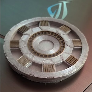 May include: A silver and gold 3D printed replica of the Arc Reactor from the Iron Man movies. The reactor has a circular shape with a central core and eight surrounding segments.