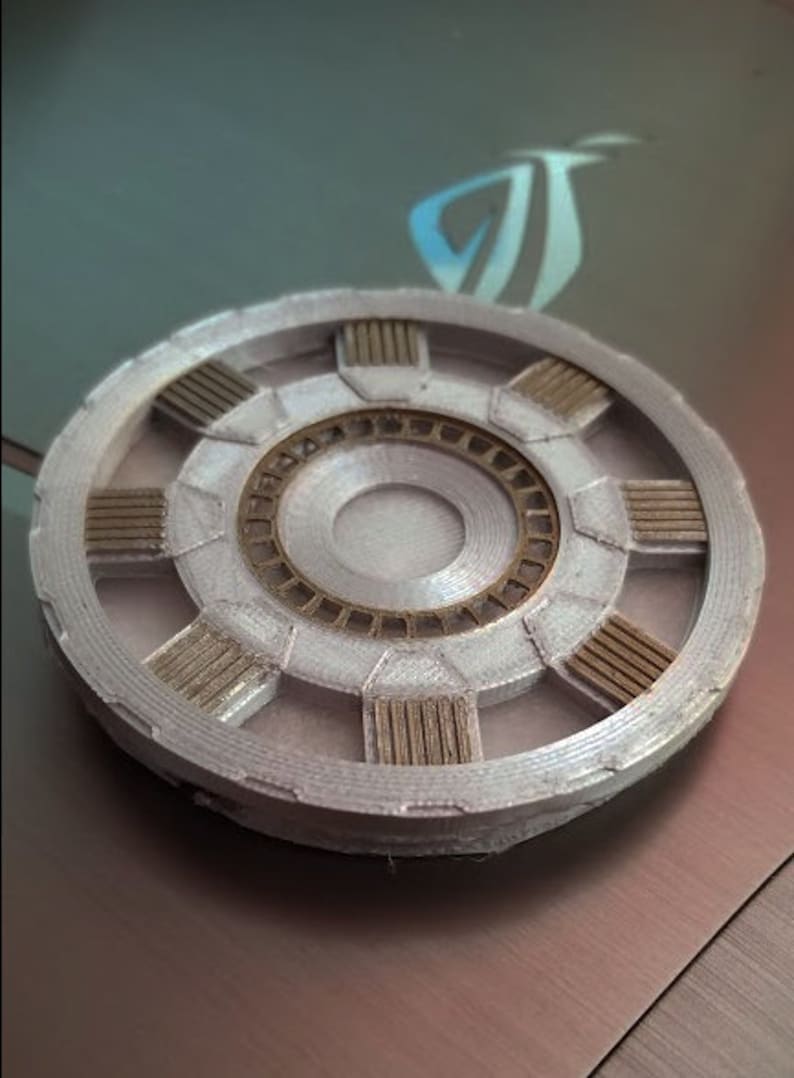 May include: A silver and gold 3D printed replica of the Arc Reactor from the Iron Man movies. The reactor has a circular shape with a central core and eight surrounding segments.
