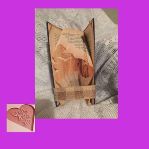 May include: A handcrafted book sculpture with pages folded to create a horse silhouette. The book is bound with a blue cover and a tan and brown band. The background is a white lace fabric.
