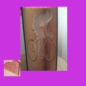 May include: A book sculpture with a cyclist design cut into the pages. The pages are a natural tan colour, with the cyclist and bicycle design visible. The book is displayed on a light-coloured surface. The text "Crafty Home" is visible.