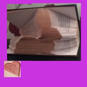 May include: A handmade book with pages folded to create a rabbit silhouette. The book is open, revealing the folded design. The pages are light brown, and the book has a dark cover. A heart-shaped tag with the words "Crafty Home" is in the corner.