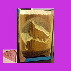 May include: A folded book art piece with a mountain silhouette carved into the pages. The book is tan and the cut-out design reveals the mountain shape. The book is set against a purple background.