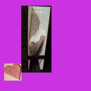 May include: A book with folded pages forming a heart shape, supported by a black stand. A small pink heart-shaped card with the words "Crafty Home" is in the lower left corner.