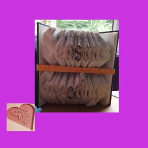 May include: A book with pages folded into a decorative design, held together by an orange band. The folded pages create a textured, layered effect. The book is open, revealing the intricate paper art. A pink heart with the words "Crafty Home" is in the corner.