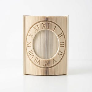 May include: A book sculpture featuring folded pages that form a circular clock face. The design displays Roman numerals from I to XII arranged in a circle, created by precisely folding the individual paper pages of the book into a clock shape.