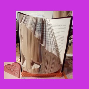 May include: A book art sculpture of a wolf, folded from the pages of a book. The book is bound in black and has a white ribbon tied around it. The sculpture is sitting on a table with an orange and white polka dot tablecloth. A pink heart-shaped sign with the text "Crafty Home" is in the lower left corner of the image.
