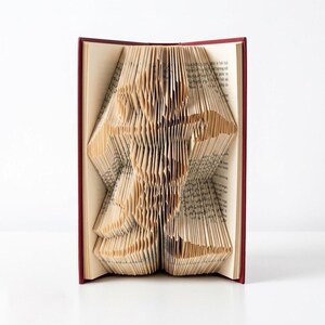 May include: A book with a maroon hard cover featuring pages folded to create a three-dimensional silhouette of a person in a running pose. The paper edges are precisely creased to form the shape, revealing printed text on the individual page surfaces.