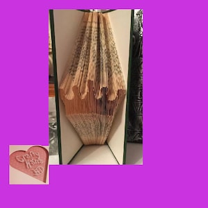 May include: A book art sculpture with pages folded into a heart shape. The book is bound in green fabric and the pages are cream coloured. The sculpture is titled "Crafty Home" in a heart-shaped frame.