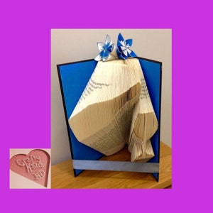 May include: A blue and white origami flower sits atop a book sculpture, which is displayed on a blue background with a black frame. The sculpture is made from the pages of a book that have been folded into a three-dimensional shape.