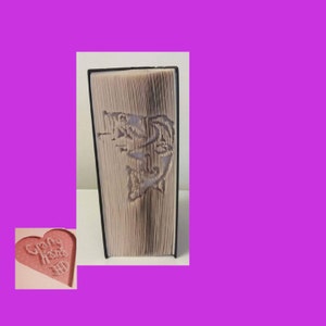 May include: A book with pages cut to reveal a fish design. The book has a black spine and the pages are light-colored. A small, pink heart-shaped object with the text "Crafty Hans" is in the lower left corner.
