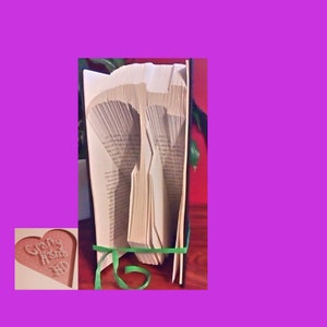 May include: A book sculpture with pages folded to create a hammer shape. The book is bound with a green ribbon. A small heart-shaped card with the words "Crafty Home" is in the lower left corner.