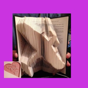 May include: A book with pages folded to create a three-dimensional star shape. The book is open, revealing the text of the pages. The craft is held by a person. A small heart-shaped card with the text "Crafty Home" is in the corner.