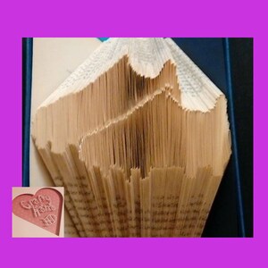Pode incluir: Uma peça de arte de livro única com um design de cordilheira esculpido nas páginas de um livro. As páginas são de cor bege quente, com as bordas cortadas para criar a silhueta da montanha. Um coração rosa com as palavras "Crafty Home" está no canto.