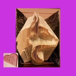 May include: A unique book art sculpture, featuring a folded book with intricate cut and folded pages. The sculpture is set against a backdrop with decorative patterns. A small heart-shaped card with the text "Crafty Home" is in the lower left corner.