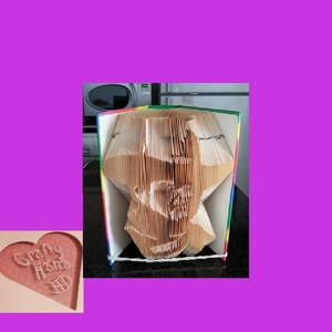 May include: A folded book sculpture with a star shape, crafted from the pages of a book. The edges of the book are multi-colored. A small, heart-shaped sign with the text "Crafty Home" is in the lower left corner.