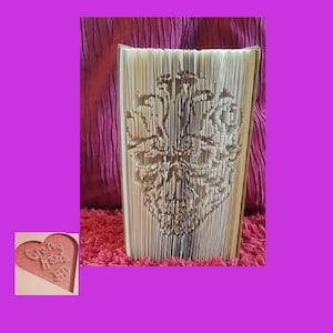 May include: An open book with folded pages forming a floral design. The book's pages are beige, set against a deep pink backdrop and a textured red surface. A small heart-shaped card with the text "Crafty Home" is in the corner. The book art is a unique decorative item.