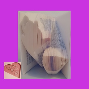 May include: A folded book with pages cut to form a heart shape. The book is open, revealing the folded pages. The text "Crafty Home" is visible on a pink heart-shaped cutout in the corner.