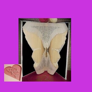 May include: A book art sculpture of a butterfly, created by folding the pages of a book. The wings are formed from the folded pages, set against a black backdrop. The artwork is framed, showcasing the intricate design.