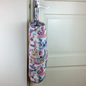 May include: A fabric bag with a floral and vintage design, hanging from a door handle. The bag features a white background with colorful illustrations of flowers, keys, and chess pieces. The bag is cylindrical and has a loop for hanging.